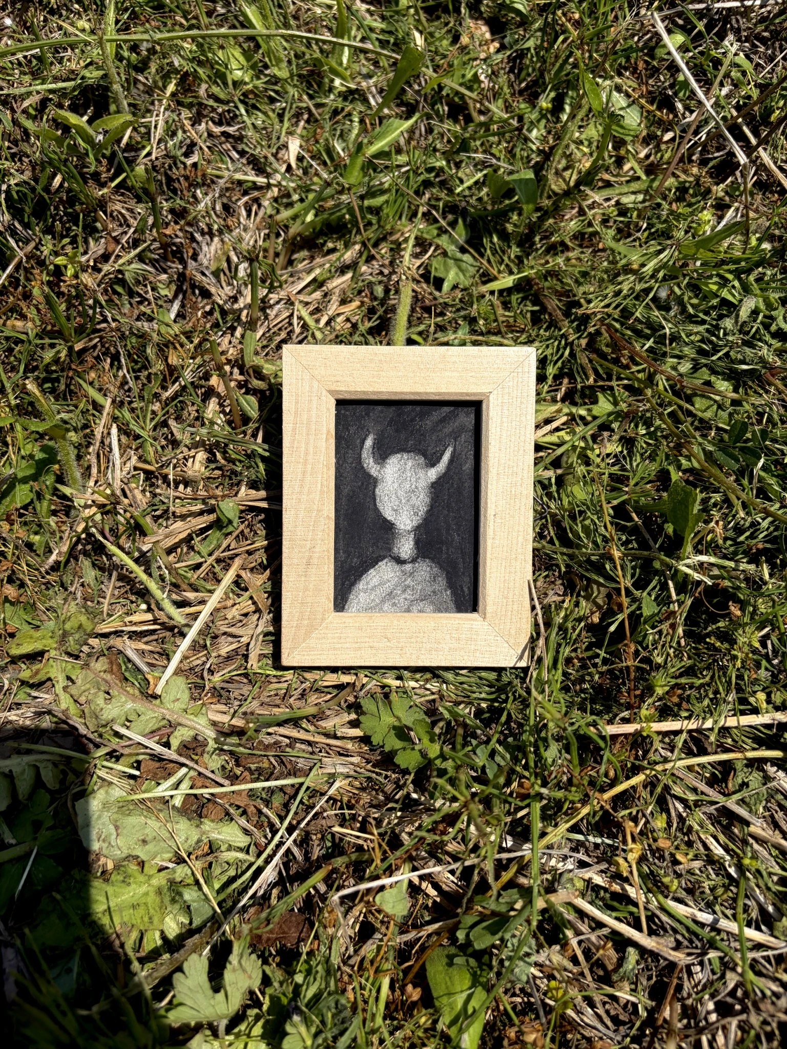 草の上に置いた木製の額に、角のある人の姿を描いた小さな作品