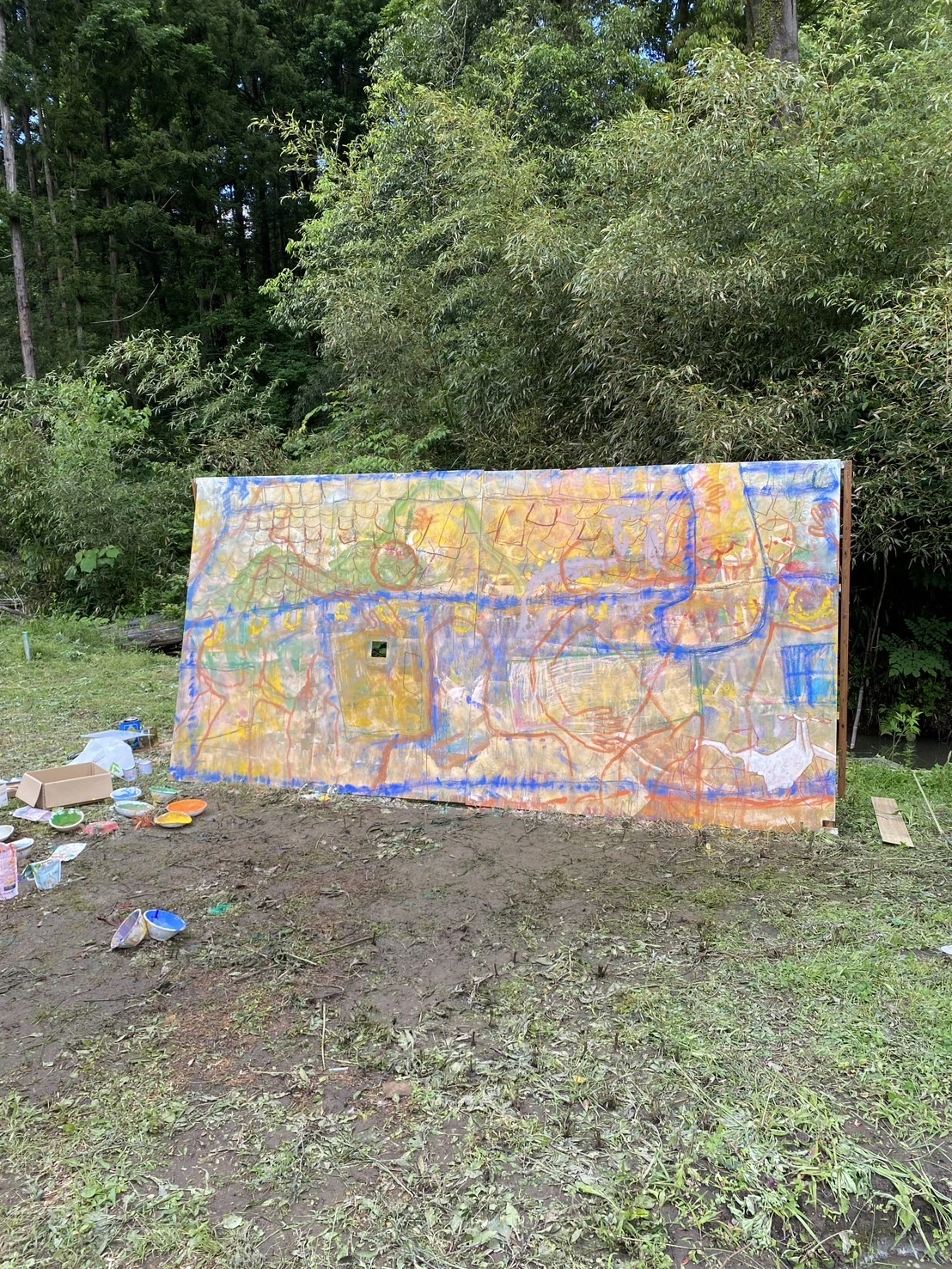 森の前の屋外に置かれた大きな絵と制作道具
