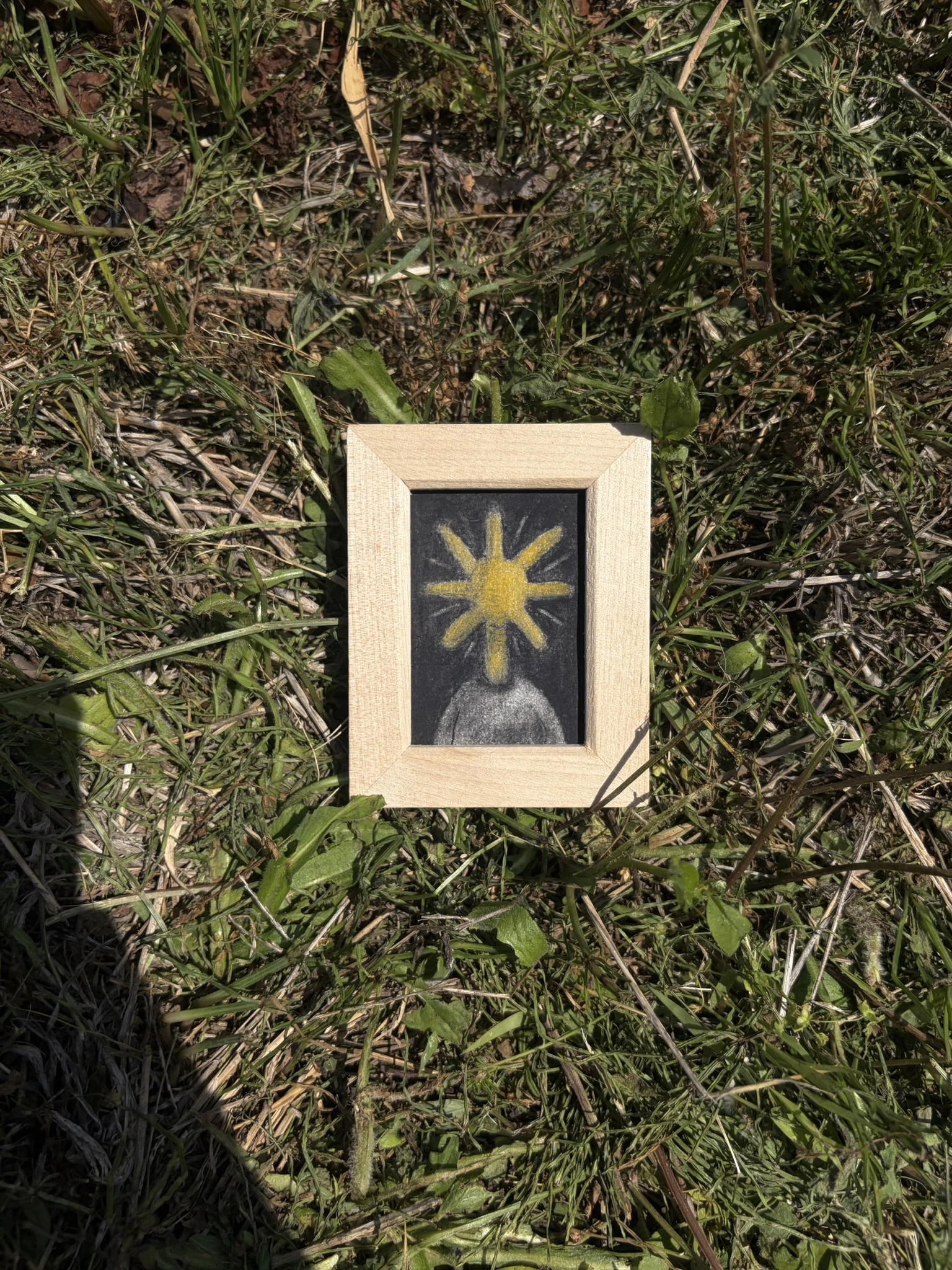草の上に置いた木製の額に、黄色い光のような頭を持つ人物を描いた小さな作品