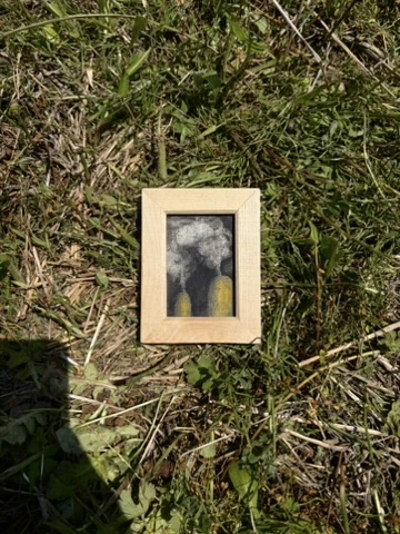 草の上に置いた木製の額に、灰色の雲と黄色い形を描いた小さな作品